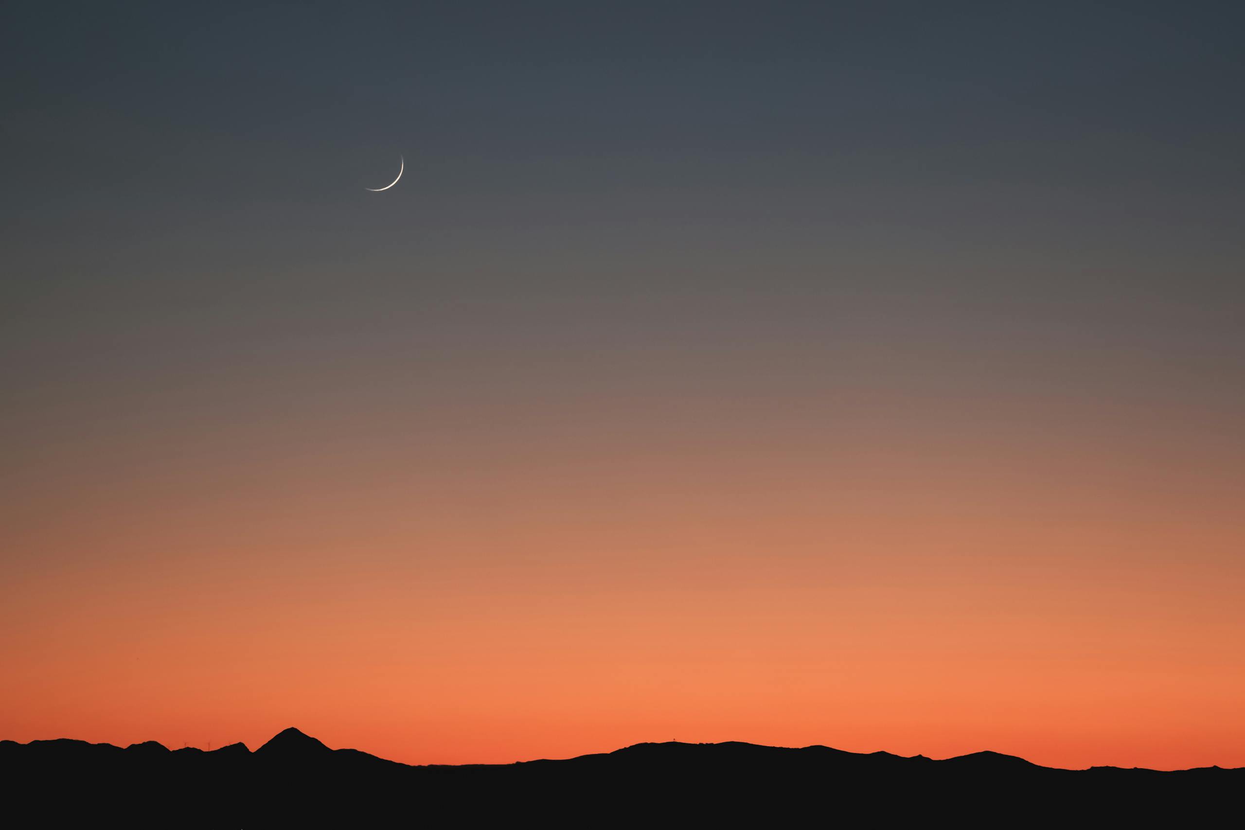 A serene sunset scene with a crescent moon over silhouetted mountains and a colorful sky.